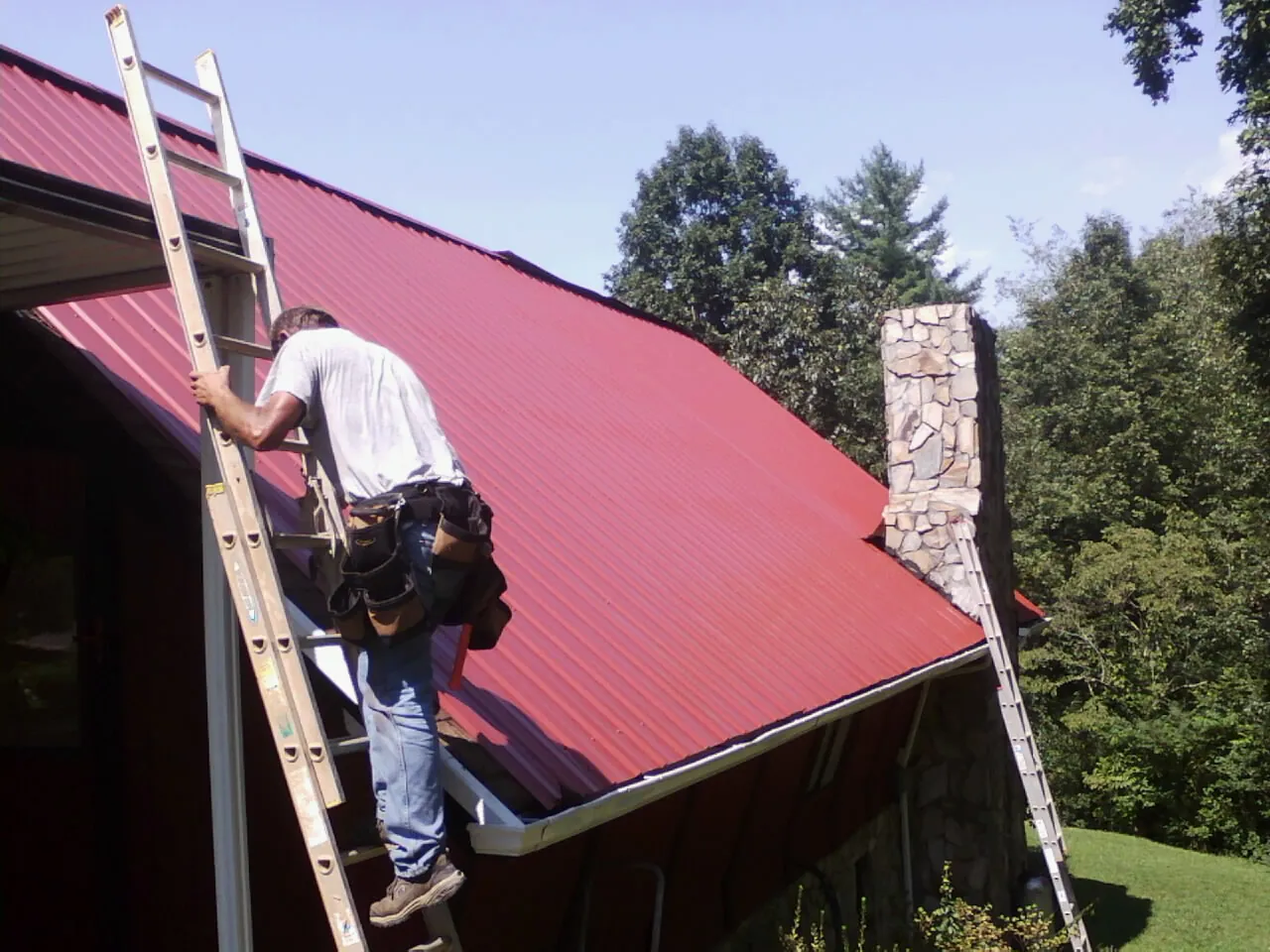 Roofer on a ladder beside a red metal roof and stone chimney — Soffit & Fascia Repair by Roofing Brothers in Merino